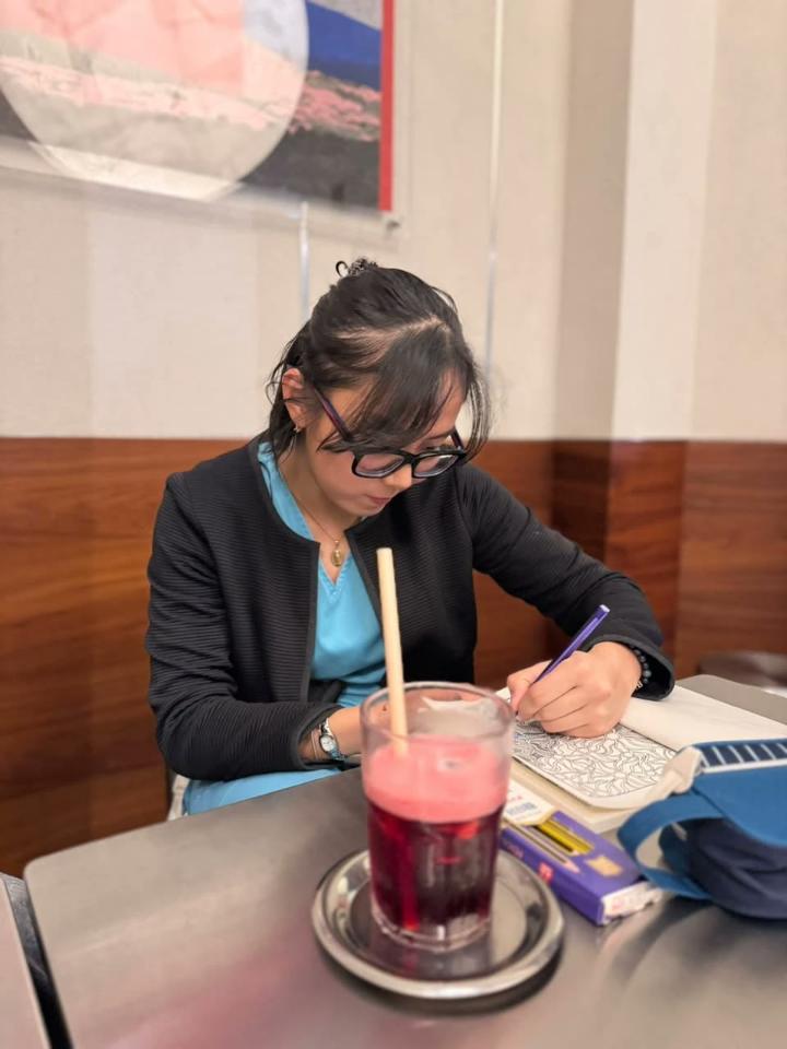 A woman in glasses works on a page with a purple pen at a café table.