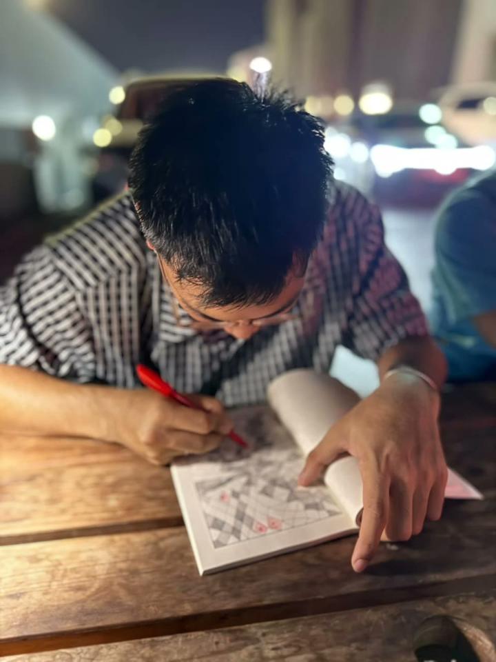 A man at an outdoor table at night fills in a pattern with a red pen.